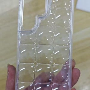 Puede incluir: Una funda transparente para tel&eacute;fono con un dise&ntilde;o de cuadr&iacute;cula y recortes para la c&aacute;mara y los botones. La funda se sostiene en una mano, mostrando su dise&ntilde;o y forma. La funda est&aacute; hecha de un material pl&aacute;stico transparente.