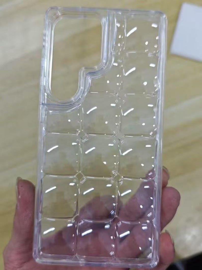Puede incluir: Una funda transparente para tel&eacute;fono con un dise&ntilde;o de cuadr&iacute;cula y recortes para la c&aacute;mara y los botones. La funda se sostiene en una mano, mostrando su dise&ntilde;o y forma. La funda est&aacute; hecha de un material pl&aacute;stico transparente.