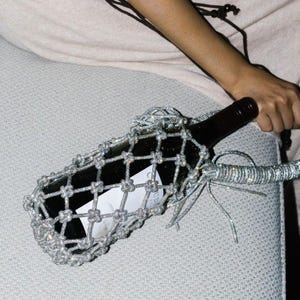 May include: A dark wine bottle is encased in a silver, diamond-like mesh netting. The netting extends into a handle, held by a person. The background is a light beige fabric.