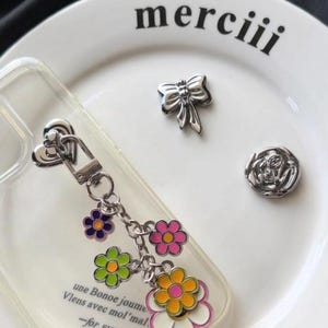 Könnte beinhalten: Eine transparente Handyhülle mit einem herzförmigen Metall-Schlüsselanhänger, der bunte Blumenanhänger in Pink, Grün, Orange und Weiß aufweist. Die Hülle trägt den Text "une Bonoe joume Viens avec mol'mal -for EVERYDAY". Enthält zwei silberne Metallanhänger: eine Schleife und eine Rose.