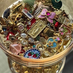 May include: A glass bowl filled with a collection of assorted metal charms, including a gold rectangular charm with a hand and star design, a pink enamel bow with a central red stone, a blue heart charm, and a small oval charm with strawberries.