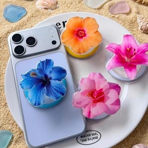 Peut inclure: Un smartphone bleu clair avec un support de téléphone en forme d'hibiscus bleu fixé au dos. Trois supports supplémentaires représentant un hibiscus orange, un lys rose et une rose rose sont disposés sur une assiette blanche avec le texte maive me.