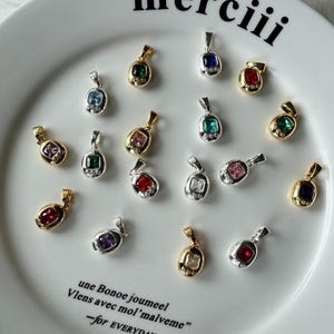 May include: A collection of small oval pendants with square-cut colored gemstones and tiny accent stones, set in gold and silver-toned metal. The pendants are arranged on a white plate featuring the text "merciii" and "une Bonoe joumeel Vlens avec mol'malveme -for EVERYDAY".