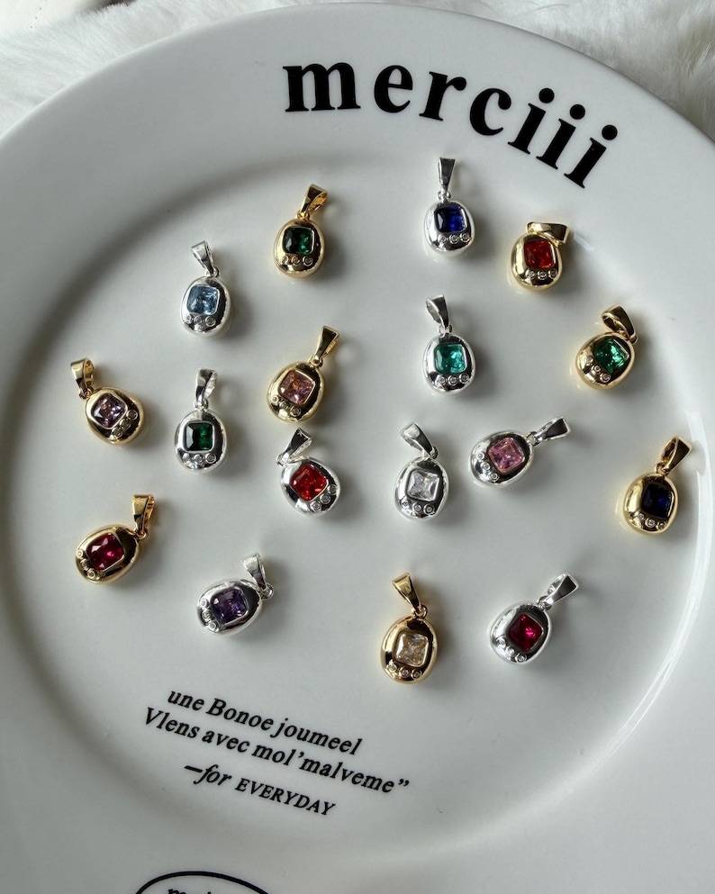 May include: A collection of small oval pendants with square-cut colored gemstones and tiny accent stones, set in gold and silver-toned metal. The pendants are arranged on a white plate featuring the text "merciii" and "une Bonoe joumeel Vlens avec mol'malveme -for EVERYDAY".