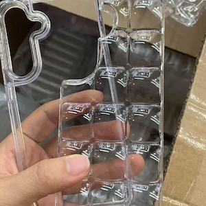 Puede incluir: Funda de tel&eacute;fono transparente y clara con un dise&ntilde;o texturizado &uacute;nico. La funda se sostiene en una mano, mostrando su forma y la forma en que captura la luz. El dise&ntilde;o presenta un patr&oacute;n de cuadr&iacute;cula de cuadrados en relieve.