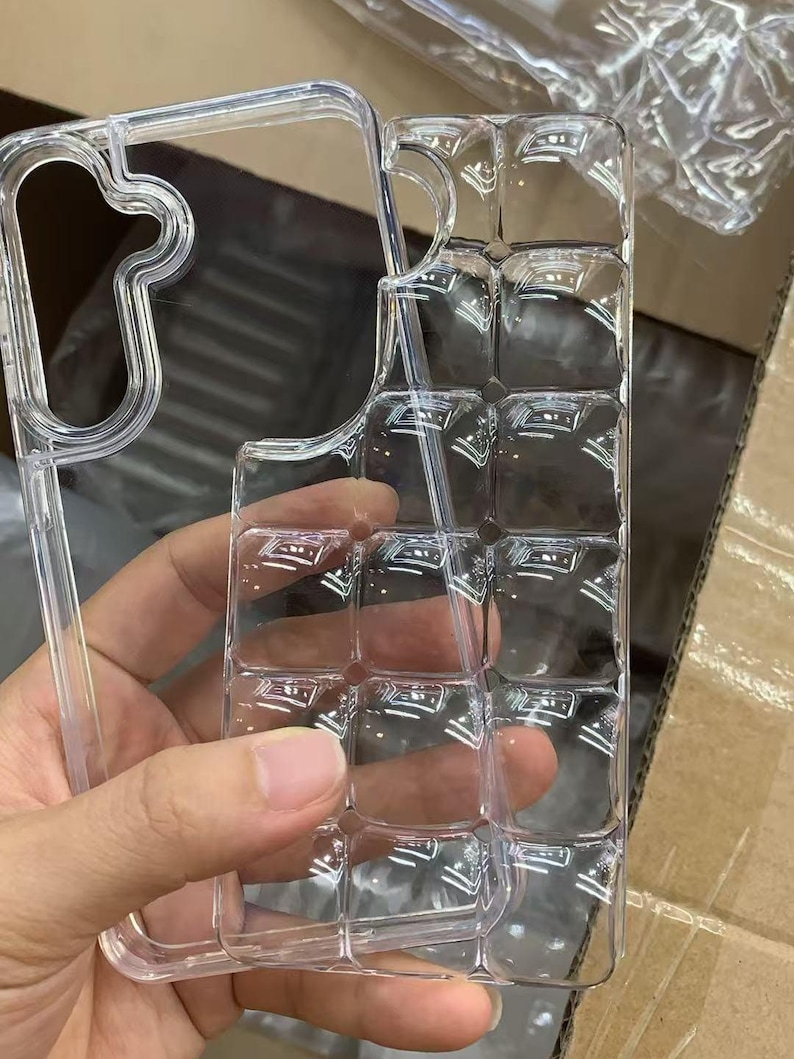 Puede incluir: Funda de tel&eacute;fono transparente y clara con un dise&ntilde;o texturizado &uacute;nico. La funda se sostiene en una mano, mostrando su forma y la forma en que captura la luz. El dise&ntilde;o presenta un patr&oacute;n de cuadr&iacute;cula de cuadrados en relieve.