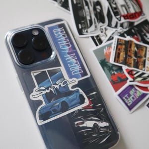 May include: Clear phone case with stickers featuring car graphics. One sticker says "DREAM CHASER" and another says "Supra" with a blue car illustration. Additional stickers with car designs are scattered around.