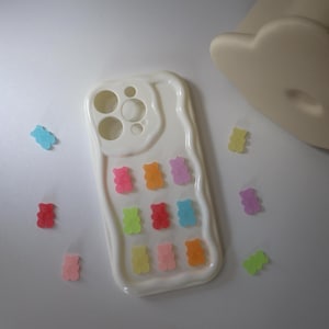 May include: White phone case with a wavy edge design, adorned with colorful gummy bear accents. The case features cutouts for the camera and buttons. Scattered around the case are additional gummy bear candies in various colors.