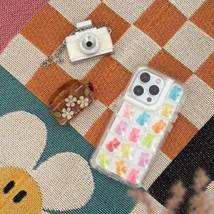 May include: A cream-colored camera pendant with a silver chain, a clear phone case filled with colorful gummy bear shapes, and a small brown floral hair clip rest on a patterned surface. The phone case has a clear border.