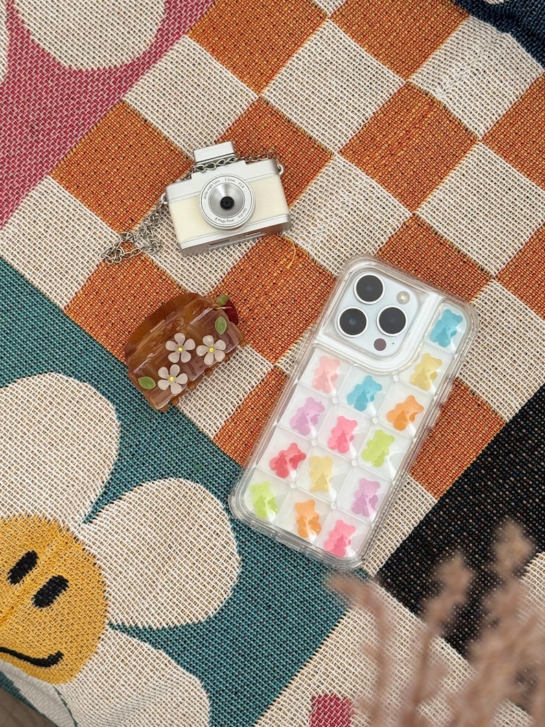 May include: A cream-colored camera pendant with a silver chain, a clear phone case filled with colorful gummy bear shapes, and a small brown floral hair clip rest on a patterned surface. The phone case has a clear border.