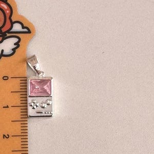 May include: A silver-toned pendant shaped like a handheld gaming console, featuring a rectangular pink gemstone screen and small circular buttons. A ruler placed alongside the pendant indicates a height of approximately 1.25 inches.