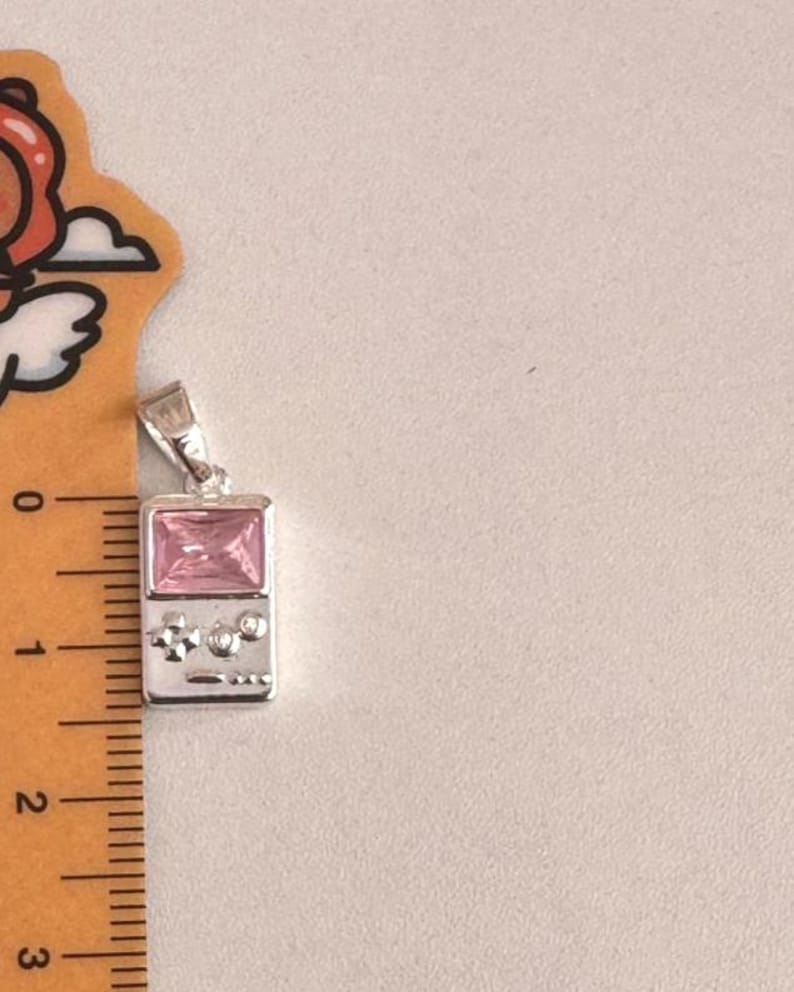 May include: A silver-toned pendant shaped like a handheld gaming console, featuring a rectangular pink gemstone screen and small circular buttons. A ruler placed alongside the pendant indicates a height of approximately 1.25 inches.