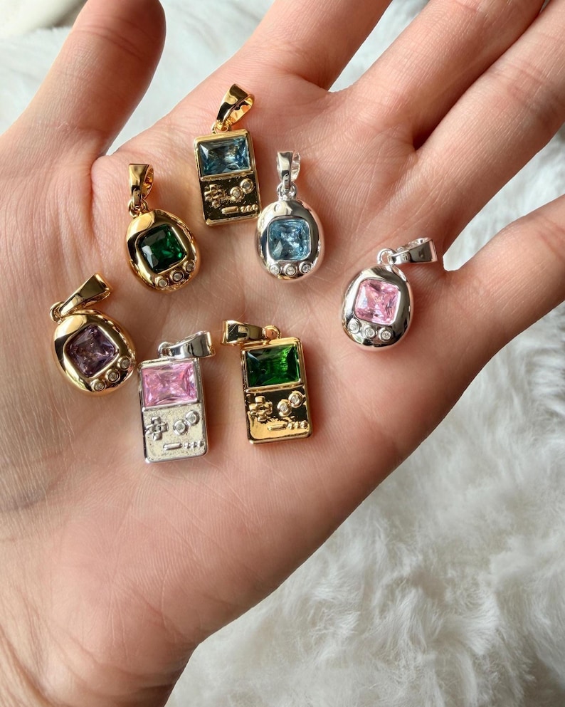 May include: A collection of six metal pendants shaped like handheld gaming consoles and oval digital pets. The pendants feature gold and silver tones with embedded rectangular or square gemstones in blue, green, pink, and purple, plus small clear accents.