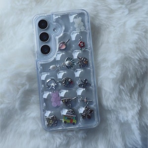 Puede incluir: Funda transparente para tel&eacute;fono con una cuadr&iacute;cula de peque&ntilde;os compartimentos, cada uno con un charm. Los charms incluyen una gema rosa, una flor, una calavera, un osito de goma y una peque&ntilde;a furgoneta. La funda est&aacute; sobre una superficie blanca y esponjosa.