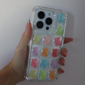 May include: Clear phone case with colorful gummy bear design. The case features a transparent design with multiple gummy bear shapes in various colors, including pink, orange, yellow, green, blue, and purple. The phone case is held in a hand.