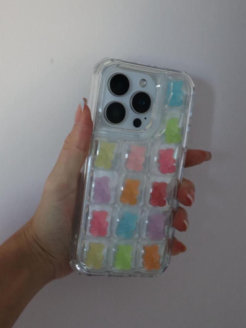 May include: Clear phone case with colorful gummy bear design. The case features a transparent design with multiple gummy bear shapes in various colors, including pink, orange, yellow, green, blue, and purple. The phone case is held in a hand.