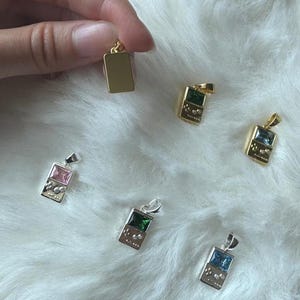 May include: A collection of rectangular pendants shaped like handheld gaming consoles, featuring metallic frames in gold and silver tones with inset colored gemstones including pink, green, and blue, each equipped with a small top loop for hanging.