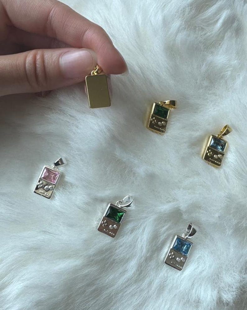 May include: A collection of rectangular pendants shaped like handheld gaming consoles, featuring metallic frames in gold and silver tones with inset colored gemstones including pink, green, and blue, each equipped with a small top loop for hanging.