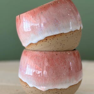 Puede incluir: Dos cuencos de cerámica apilados con un esmalte rosa y blanco. Los cuencos tienen un exterior marrón moteado.