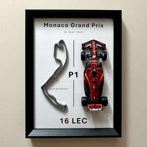 Peut inclure: Une impression encadrée d'un circuit de course automobile avec une voiture de course rouge sur fond blanc. Le texte "Monaco Grand Prix 26 MAI 2024" est en haut de l'impression. Le numéro "16 LEC" est en bas de l'impression.