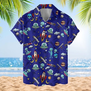 May include: A short-sleeved button-down shirt in deep blue featuring an all-over space-themed print. The pattern includes light green aliens, flying saucers, red and white rockets, yellow crescent moons, telescopes, and various planets.