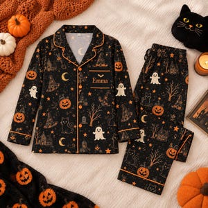 May include: A black long-sleeve pajama set with orange piping, featuring a repeating Halloween pattern of pumpkins, ghosts, cats, witch hats, and moons. The chest pocket is embroidered with the name Emma in a gold-colored script font.