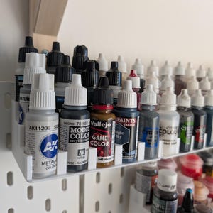 May include: A selection of acrylic paint bottles in different colours, arranged on a white shelf. The bottles have black and white caps, with labels showing brand names and colour names. Some bottles are labelled "Metallic Colour" and "Game Colour".
