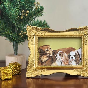 May include: A gold framed painting of three dogs, including a golden retriever and two Cavalier King Charles Spaniels. The frame has decorative detailing. A small Christmas tree and gold gift boxes are in the background.