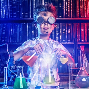May include: A young person wearing goggles and a white shirt is conducting a science experiment in a laboratory setting. They are holding beakers filled with colorful liquids and stirring them with a glass rod. The lab is filled with various beakers, flasks, and other scientific equipment. The background features a bookshelf filled with books.