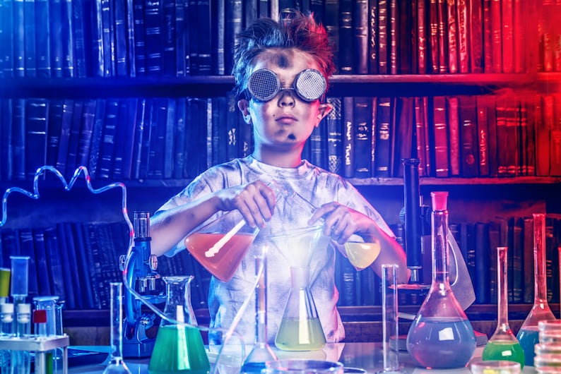 May include: A young person wearing goggles and a white shirt is conducting a science experiment in a laboratory setting. They are holding beakers filled with colorful liquids and stirring them with a glass rod. The lab is filled with various beakers, flasks, and other scientific equipment. The background features a bookshelf filled with books.