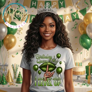 May include: A woman wearing a silver crown and a gray t-shirt with the text "Birthday Queen" and a green lip design. The shirt also says "I don't get older I LEVEL UP". The background includes balloons, a cake, and a banner that says "HAPPY".