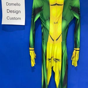 Op de afbeelding: Groen en geel spandex pak met een comic-stijl print. Het pak heeft een hoge hals en lange mouwen. De tekst "Domello Design Custom" is zichtbaar op een wit bord.