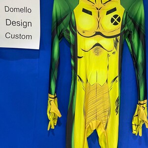 Op de afbeelding: Een gele en groene spandex jumpsuit met een zwarte X op de borst en zwarte lijnen over de hele stof. De jumpsuit heeft een hoge hals en lange mouwen.