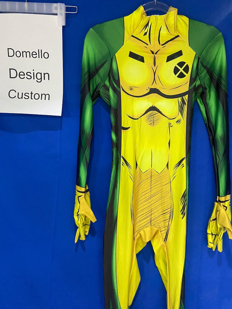 Op de afbeelding: Een gele en groene spandex jumpsuit met een zwarte X op de borst en zwarte lijnen over de hele stof. De jumpsuit heeft een hoge hals en lange mouwen.
