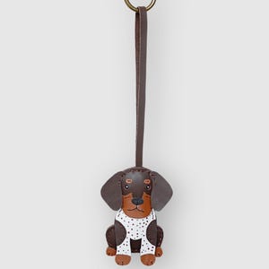May include: A leather dachshund dog keyring in brown and black, wearing a white polka dot shirt. It has a brown leather strap and a gold-coloured ring. The text "SC CRAFT" is printed at the bottom.