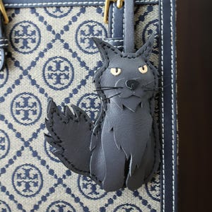 May include: A black leather cat keyring with stitched details. The keyring is attached to a black leather strap with a gold buckle. The cat is sitting with its tail curled around its legs. The keyring is hanging from a blue and white patterned handbag.