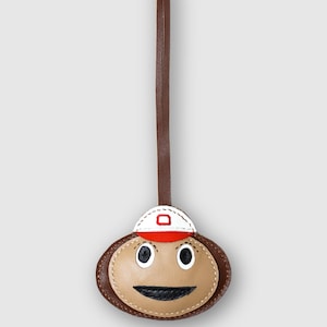 May include: A brown leather luggage tag shaped like a face wearing a white and red baseball cap with a red letter O on the front. The face has black eyes and a black mouth. A long brown leather strap extends from the top. The text SC CRAFT is visible.