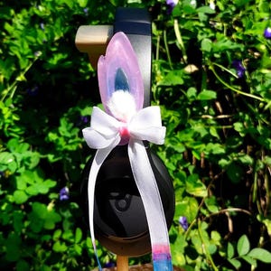 Puede incluir: Auriculares negros con una diadema de orejas de conejo rosa y blanca. La diadema tiene un lazo blanco y un amuleto iridiscente rosa y azul.