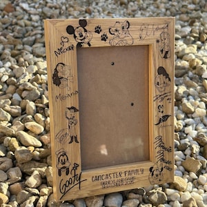 May include: A wooden picture frame featuring engraved Disney characters such as Mickey Mouse, Simba, and Goofy. The frame also includes the text "Lancaster Family Disneyland Paris September 2025."