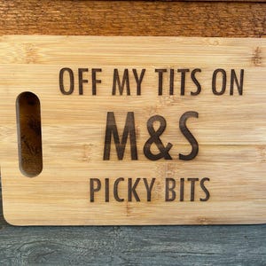 Könnte beinhalten: Rechteckiges Schneidebrett aus hellem Bambus mit Griffaussparung. Der Text "OFF MY TITS ON M&S PICKY BITS" ist in dunkler Schrift eingraviert. Das Brett ist aus Holz gefertigt.