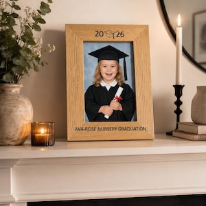 May include: A light oak wood picture frame featuring the engraved text 20 26 with a graduation cap icon at the top and AVA-ROSE NURSERY GRADUATION at the bottom. The frame holds a photograph of a child wearing a black graduation cap and gown.