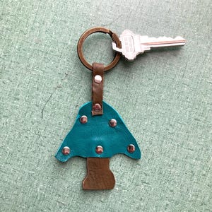 May include: Teal leather mushroom keychain with brown leather stem and silver rivets. The keychain has a brass key ring with a silver key attached.