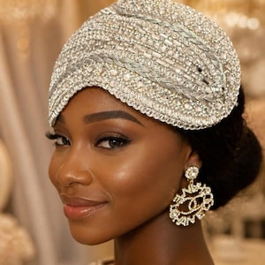 May include: A silver headpiece adorned with intricate beadwork, clear crystals, and braided metallic trim. The headpiece features a dense, textured pattern of shimmering embellishments covering the entire surface, paired with large, circular crystal earrings.