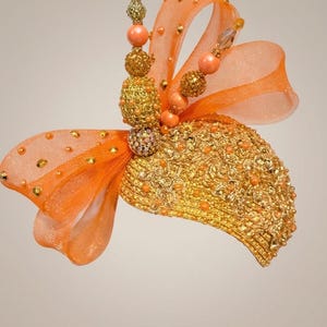 Könnte beinhalten: Ein goldfarbener Kopfschmuck mit einer strukturierten, metallischen Basis, die mit kleinen orangefarbenen Perlen und goldener Stickerei verziert ist. Er ist mit großen orangefarbenen Organzaschleifen, goldfarbenen Perlensträngen und klaren Kristallen akzentuiert.