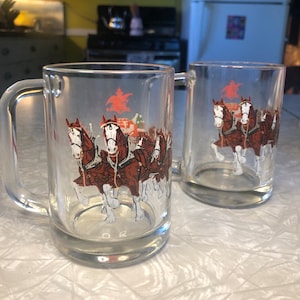 May include: Two clear glass beer mugs with a Budweiser Clydesdale horse and wagon graphic. The mugs are on a white surface.