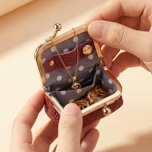 May include: A small, burgundy leather jewellery case with a gold clasp, open to reveal a gold necklace and rings. The interior is lined with grey fabric and polka dots. The case is held open by two hands.