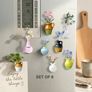 May include: A collection of eight miniature vases, each with artificial flowers, attached to a fridge. The vases are in various colours, including pink, yellow, blue, and white. A card with the text "Enjoy the little things" is also attached to the fridge.