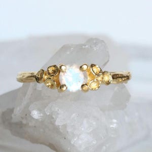 May include: A gold ring with a round, iridescent gemstone surrounded by small, textured gold accents. The ring is set on a white, textured surface.
