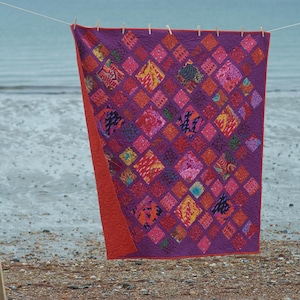 Op de afbeelding: Een kleurrijk quilt met een ruitpatroon in tinten roze, rood, oranje en paars. De quilt hangt aan een waslijn tegen een achtergrond van een zandstrand en blauwe zee.