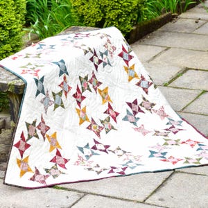 May include: A white quilt with a pattern of colorful stars. The quilt is made with a variety of fabrics, including floral prints and solid colors. The quilt is folded and draped over a wooden bench.